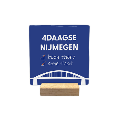 4daagse Nijmegen - Decoratie tegel - Formaat: - Op deze decoratie tegel staat bovenaan de tekst "4daagse Nijmegen" in witte hoofdletters. Daaronder staan twee regels met de tekst "been there" en "done that", waarbij voor elke regel een wit selectievakje met een oranje vinkje is geplaatst. Onderaan het ontwerp is een gestileerde witte afbeelding van een brug zichtbaar. Het geheel is geplaatst op een donkerblauwe achtergrond. - Snelwenskaart
