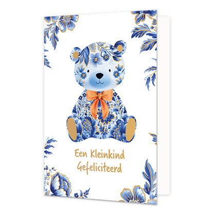 Een kleinkind gefeliciteerd - Formaat: Normaal - Vorm: Staand - Geboortekaart - Op deze wenskaart staat centraal een zittende teddybeer afgebeeld die volledig is gedecoreerd met blauw-witte bloemmotieven in Delfts blauwe stijl, gecombineerd met effen gouden binnenoren en voetzolen. De beer draagt een opvallende, oranje gestrikte sjaal om de nek. De hoeken van de kaart zijn versierd met bijpassende blauwe bloemen en bladeren met gouden randen tegen een witte achtergrond. Onderaan de illustratie staat in gou