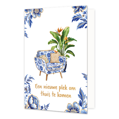 Een nieuwe plek om thuis te komen - Formaat: Normaal - Vorm: Staand - Woningkaart - Op deze wenskaart staat centraal een fauteuil afgebeeld die is bekleed met een blauw-wit bloemenpatroon in Delfts blauwe stijl, voorzien van twee kussens met een goudkleurig dessin en houten pootjes. Direct achter de stoel staat een grote, groene Strelitzia-plant (paradijsvogelbloem) met oranje-blauwe bloemen en goudkleurige randen langs de bladeren. De hoeken van de kaart zijn gedecoreerd met bijpassende blauwe bloemen en 