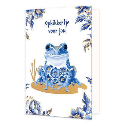 Opkikkertje voor jou - Formaat: Normaal - Vorm: Staand - Opkikker kaart - Op deze wenskaart staat centraal een kikker afgebeeld die is gedecoreerd met blauw-witte bloemmotieven in Delfts blauwe stijl en goudkleurige stippen op de poten en rug. De kikker zit op een goudkleurig vlak met daarnaast twee goudkleurige rietstengels, terwijl de hoeken van de kaart zijn versierd met bijpassende blauwe bloemen en bladeren met gouden randen tegen een witte achtergrond. Bovenaan de illustratie staat in blauwe, handges