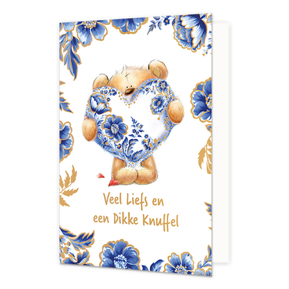  Op deze wenskaart staat centraal een pluche teddybeer afgebeeld die een groot hart vasthoudt dat volledig is versierd met blauw-witte bloemmotieven in Delfts blauwe stijl en goudkleurige stippen. De randen en hoeken van de kaart zijn rijkelijk gedecoreerd met bijpassende blauwe bloemen en bladeren met gouden accenten tegen een witte achtergrond. Onderaan de illustratie staat in goudkleurige, handgeschreven letters de tekst "Veel Liefs en een Dikke Knuffel". 