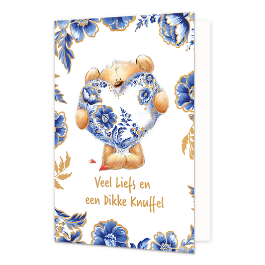  Op deze wenskaart staat centraal een pluche teddybeer afgebeeld die een groot hart vasthoudt dat volledig is versierd met blauw-witte bloemmotieven in Delfts blauwe stijl en goudkleurige stippen. De randen en hoeken van de kaart zijn rijkelijk gedecoreerd met bijpassende blauwe bloemen en bladeren met gouden accenten tegen een witte achtergrond. Onderaan de illustratie staat in goudkleurige, handgeschreven letters de tekst "Veel Liefs en een Dikke Knuffel". 