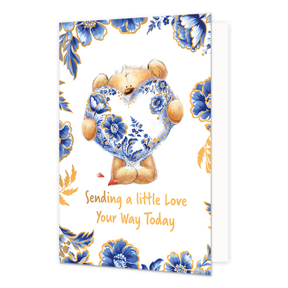 Sending a little love your way today - Formaat: Normaal - Vorm: Staand - Denken aan kaart - Op deze wenskaart staat centraal een schattige, zittende teddybeer afgebeeld, die een groot hart vasthoudt dat versierd is met gedetailleerde blauw-witte Delfts Blauwe bloemmotieven en gouden accenten. De beer zelf heeft een lichtbruine kleur en kijkt met grote, vriendelijke ogen naar voren, met kleine rode hartjes bij zijn poten. De randen en hoeken van de kaart zijn rijkelijk versierd met bijpassende grote blauwe 