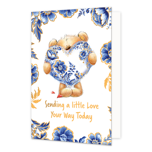 Sending a little love your way today - Formaat: Normaal - Vorm: Staand - Denken aan kaart - Op deze wenskaart staat centraal een schattige, zittende teddybeer afgebeeld, die een groot hart vasthoudt dat versierd is met gedetailleerde blauw-witte Delfts Blauwe bloemmotieven en gouden accenten. De beer zelf heeft een lichtbruine kleur en kijkt met grote, vriendelijke ogen naar voren, met kleine rode hartjes bij zijn poten. De randen en hoeken van de kaart zijn rijkelijk versierd met bijpassende grote blauwe 