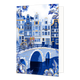 Amsterdam - Formaat: Standaard - Vorm: Staand - Blanco kaart - Op deze wenskaart is een sfeervol Nederlands stadsgezicht aan een gracht afgebeeld met karakteristieke grachtenpanden in blauwe en witte tinten met goudkleurige accenten. Op de stenen boogbrug over het water staat een fiets die is gedecoreerd met Delfts blauwe patronen en een mandje met rode tulpen aan het stuur. De hoeken en randen van de kaart zijn rijkelijk versierd met grote, gedetailleerde blauwe bloemen en bladeren met goudkleurige randen