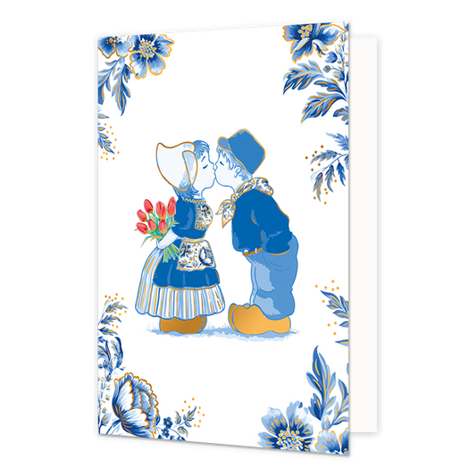 Kussend paar - Formaat: Normaal - Vorm: Staand - Blanco kaart - Op deze wenskaart staat een illustratie van een kussend jongetje en meisje in traditionele Nederlandse klederdracht, waarbij het meisje een boeket rode tulpen achter haar rug vasthoudt. De kinderen dragen blauwe kleding en goudkleurige klompen, wat aansluit bij de Delfts blauwe stijl van de omlijstende decoratie. De vier hoeken van de kaart zijn rijkelijk versierd met grote blauwe bloemen, bladeren en goudkleurige accenten tegen een witte acht