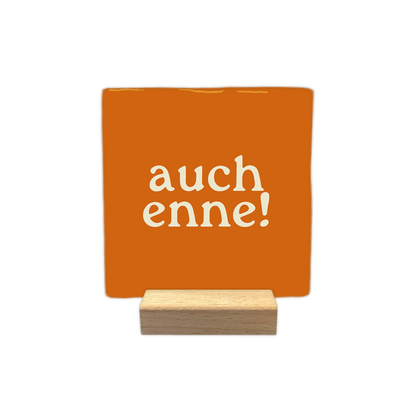 Auch enne! - Decoratie tegel - Formaat: - Op deze decoratie tegel staat de tekst "auch enne!" in crème kleurige letters. De tekst is verdeeld over twee regels op een oranje achtergrond. Het gebruikte lettertype is speels en heeft schreven. - Snelwenskaart