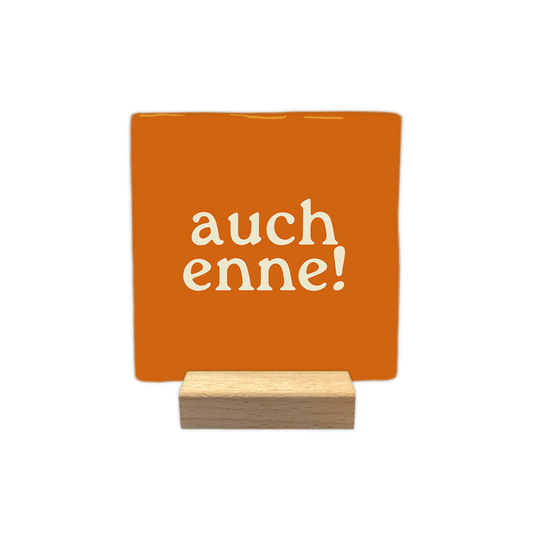 Auch enne! - Decoratie tegel - Formaat: - Op deze decoratie tegel staat de tekst "auch enne!" in crème kleurige letters. De tekst is verdeeld over twee regels op een oranje achtergrond. Het gebruikte lettertype is speels en heeft schreven. - Snelwenskaart