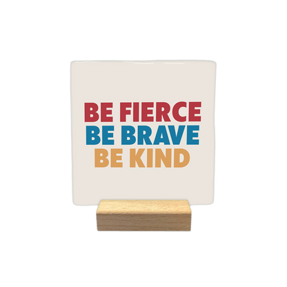 Be fierce be brave be kind - Decoratie tegel - Formaat: - Op deze decoratietegel staat de tekst "BE FIERCE", "BE BRAVE" en "BE KIND" onder elkaar in hoofdletters. De woorden zijn in drie verschillende kleuren uitgevoerd: de bovenste regel is rood, de middelste is blauw en de onderste is beige. De tekst is gecentreerd op een lichtbeige achtergrond. - Snelwenskaart