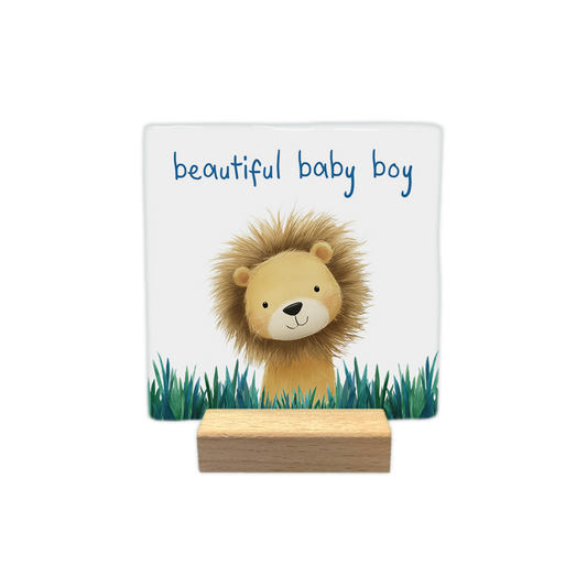 Beautiful baby boy - Decoratie tegel - Formaat: - Op deze decoratie tegel staat een illustratie van een schattig leeuwtje met warrige manen dat boven groen gras uitkijkt tegen een witte achtergrond. Het leeuwtje kijkt vriendelijk naar voren en heeft zachte gelaatstrekken. Bovenaan de illustratie staat in een blauw lettertype de tekst "beautiful baby boy" geschreven. - Snelwenskaart