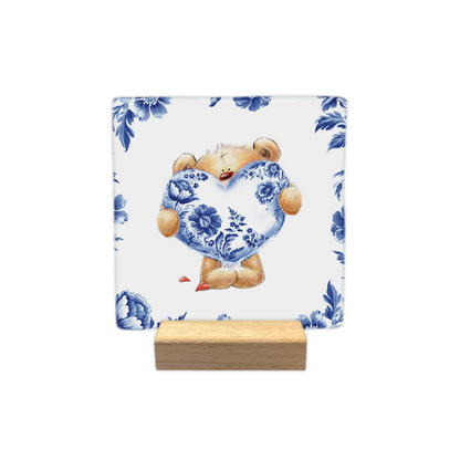 Beertje met Delfts hart - Decoratie tegel - Formaat: - Op deze decoratie tegel staat een knuffelbeer afgebeeld die een groot hart vasthoudt dat gedecoreerd is met een Delfts blauw bloemmotief. De beer is omringd door een rand van dezelfde Delfts blauwe bloemen op een witte achtergrond. Naast de voeten van de beer liggen twee kleine, rode hartjes. - Snelwenskaart