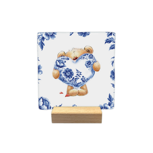 Beertje met Delfts hart - Decoratie tegel - Formaat: - Op deze decoratie tegel staat een knuffelbeer afgebeeld die een groot hart vasthoudt dat gedecoreerd is met een Delfts blauw bloemmotief. De beer is omringd door een rand van dezelfde Delfts blauwe bloemen op een witte achtergrond. Naast de voeten van de beer liggen twee kleine, rode hartjes. - Snelwenskaart