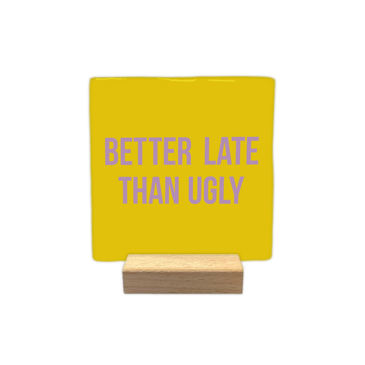 Better late than ugly - Decoratie tegel - Formaat: - Op deze decoratietegel staat in het midden de Engelse tekst "BETTER LATE THAN UGLY". De tegel zelf is okergeel en vierkant, en staat in een houten standaard. De tekst is verdeeld over twee regels en is geschreven in strakke, paarse hoofdletters. - Snelwenskaart