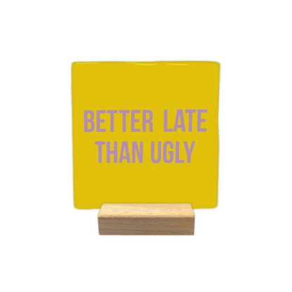 Better late than ugly - Decoratie tegel - Formaat: - Op deze decoratietegel staat in het midden de Engelse tekst "BETTER LATE THAN UGLY". De tegel zelf is okergeel en vierkant, en staat in een houten standaard. De tekst is verdeeld over twee regels en is geschreven in strakke, paarse hoofdletters. - Snelwenskaart