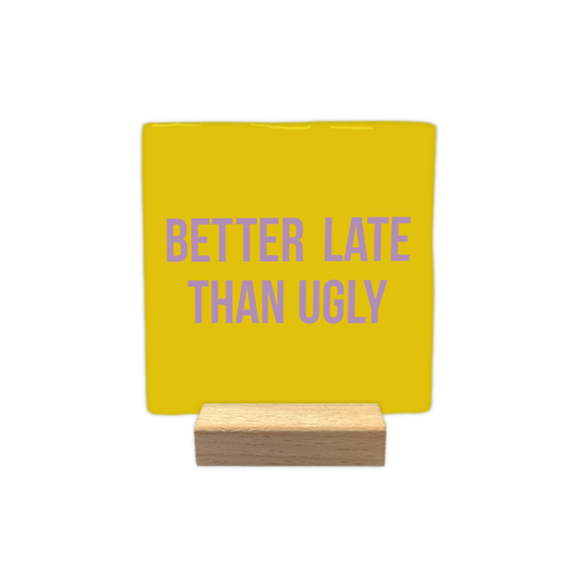 Better late than ugly - Decoratie tegel - Formaat: - Op deze decoratietegel staat in het midden de Engelse tekst "BETTER LATE THAN UGLY". De tegel zelf is okergeel en vierkant, en staat in een houten standaard. De tekst is verdeeld over twee regels en is geschreven in strakke, paarse hoofdletters. - Snelwenskaart