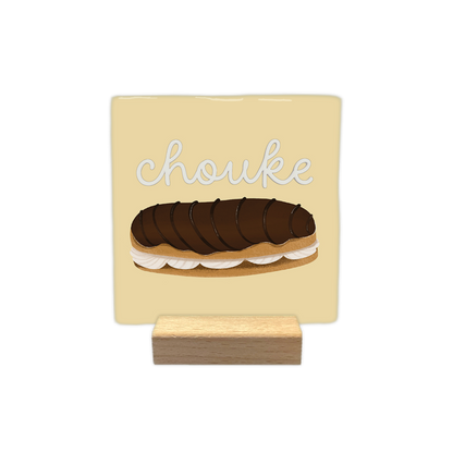 Chouke - Decoratie tegel - Formaat: - Op deze decoratie tegel staat bovenin de tekst "chouke" in witte, zwierige letters geschreven. Daaronder is een illustratie afgebeeld van een eclair met een chocoladeglazuurlaag en een vulling van slagroom. De achtergrond van het ontwerp heeft een zachtgele kleur. - Snelwenskaart
