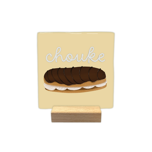 Chouke - Decoratie tegel - Formaat: - Op deze decoratie tegel staat bovenin de tekst "chouke" in witte, zwierige letters geschreven. Daaronder is een illustratie afgebeeld van een eclair met een chocoladeglazuurlaag en een vulling van slagroom. De achtergrond van het ontwerp heeft een zachtgele kleur. - Snelwenskaart