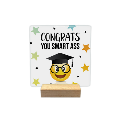Congrats you smart ass - Decoratie tegel - Formaat: - Op deze decoratie tegel staat een gele smiley met ronde bril en een afstudeerhoed op. Op de achtergrond staan gekleurde sterren en zwarte stippen. Boven de smiley staat in het zwart de tekst 'Congrats you smart ass'. - Snelwenskaart