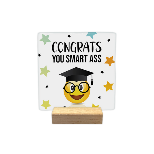 Congrats you smart ass - Decoratie tegel - Formaat: - Op deze decoratie tegel staat een gele smiley met ronde bril en een afstudeerhoed op. Op de achtergrond staan gekleurde sterren en zwarte stippen. Boven de smiley staat in het zwart de tekst 'Congrats you smart ass'. - Snelwenskaart