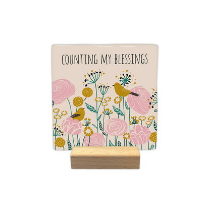 Counting my blessings - Decoratie tegel - Formaat: - Op deze decoratietegel staat bovenaan de tekst "counting my blessings" in zwarte hoofdletters tegen een crèmekleurige achtergrond. Daaronder is een levendig botanisch patroon afgebeeld met gestileerde bloemen in roze, mosterdgeel en zeegroen. Te midden van de bloemen zijn twee mosterdgele vogeltjes te zien, die op verschillende bloemen rusten. De algemene stijl van de illustratie is modern en vintage, met gedetailleerde vormen en patronen. - Snelwenskaar