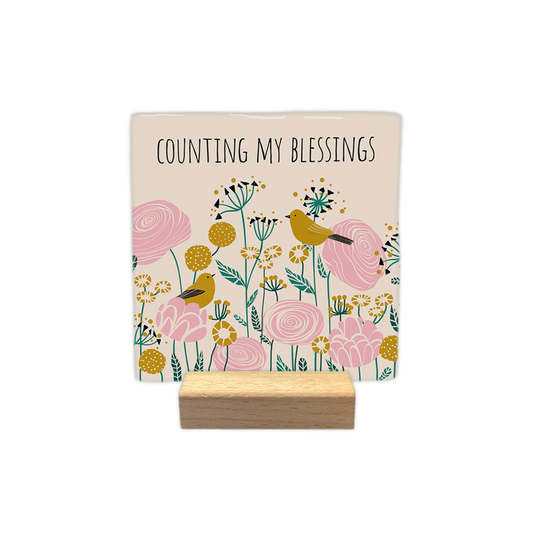 Counting my blessings - Decoratie tegel - Formaat: - Op deze decoratietegel staat bovenaan de tekst "counting my blessings" in zwarte hoofdletters tegen een crèmekleurige achtergrond. Daaronder is een levendig botanisch patroon afgebeeld met gestileerde bloemen in roze, mosterdgeel en zeegroen. Te midden van de bloemen zijn twee mosterdgele vogeltjes te zien, die op verschillende bloemen rusten. De algemene stijl van de illustratie is modern en vintage, met gedetailleerde vormen en patronen. - Snelwenskaar