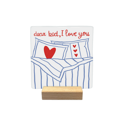 Dear bed, I love you - Decoratie tegel - Formaat: - Op deze decoratietegel staat bovenaan de tekst "dear bed, I love you" in een handgeschreven rood lettertype. Daaronder is een illustratie afgebeeld van een opgemaakt bed met blauw-witte strepen, voorzien van kussens die zijn versierd met rode hartjes. Het ontwerp is gecentreerd op een witte achtergrond. - Snelwenskaart