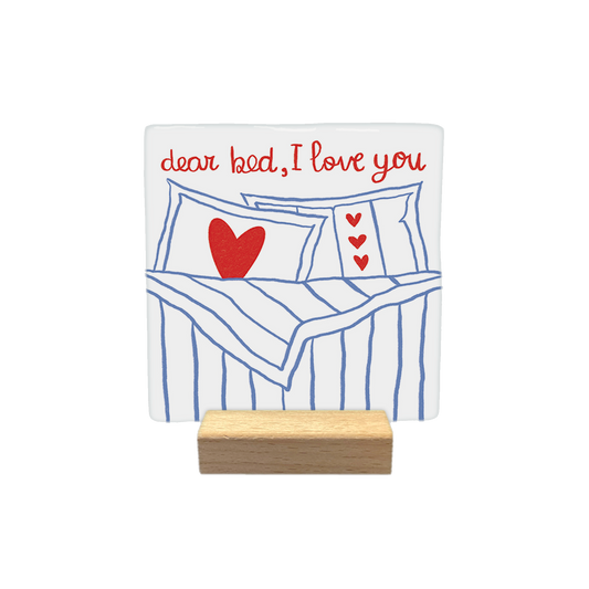 Dear bed, I love you - Decoratie tegel - Formaat: - Op deze decoratietegel staat bovenaan de tekst "dear bed, I love you" in een handgeschreven rood lettertype. Daaronder is een illustratie afgebeeld van een opgemaakt bed met blauw-witte strepen, voorzien van kussens die zijn versierd met rode hartjes. Het ontwerp is gecentreerd op een witte achtergrond. - Snelwenskaart