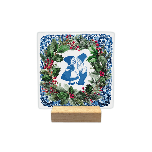 Delfts blauw kussend koppel - Decoratie tegel - Formaat: - Op deze decoratie tegel staat een afbeelding van een zoenend paartje in traditionele blauw-witte kleding. Het tafereel is omringd door een groene kerstkrans met rode bessen en dennentakken. De randen van de tegel hebben een blauw-wit Delfts blauw patroon. - Snelwenskaart