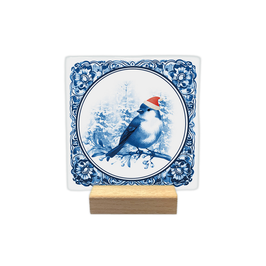 Delfts blauwe kerst vogel - Decoratie tegel - Formaat: - Op deze decoratie tegel staat een blauw vogeltje met een rood kerstmutsje op dat op een tak zit. De achtergrond toont een besneeuwd bos in blauwtinten binnen een ronde omlijsting. De randen van de tegel zijn versierd met een traditioneel blauw-wit Delfts blauw patroon. - Snelwenskaart