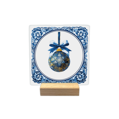 Delfts blauwe kerstbal - Decoratie tegel - Formaat: - Op deze decoratie tegel staat een blauwe kerstbal afgebeeld met gouden accenten en een witte sneeuwvlok in het midden. De bal hangt aan een blauw lint met een gestrikte strik eraan. Het centrale gedeelte bevindt zich binnen een ronde omlijsting op een witte achtergrond. De randen van de tegel zijn versierd met een klassiek blauw-wit Delfts blauw patroon. - Snelwenskaart