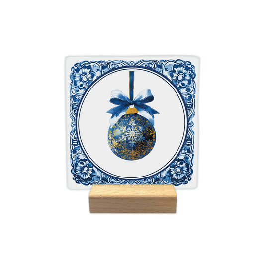 Delfts blauwe kerstbal - Decoratie tegel - Formaat: - Op deze decoratie tegel staat een blauwe kerstbal afgebeeld met gouden accenten en een witte sneeuwvlok in het midden. De bal hangt aan een blauw lint met een gestrikte strik eraan. Het centrale gedeelte bevindt zich binnen een ronde omlijsting op een witte achtergrond. De randen van de tegel zijn versierd met een klassiek blauw-wit Delfts blauw patroon. - Snelwenskaart