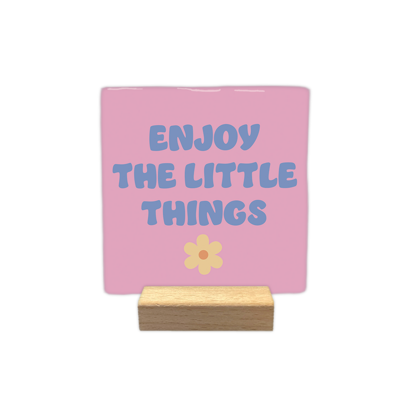 Enjoy the little things - Decoratie tegel - Formaat: - Op deze decoratietegel staat in het midden de Engelse tekst "ENJOY THE LITTLE THINGS". De tegel zelf is roze en vierkant, en staat in een houten standaard. De tekst is verdeeld over drie regels en geschreven in een speels, blauwachtig paars lettertype in hoofdletters, met daaronder een kleine, lichtgele bloem met een oranje rondje. - Snelwenskaart