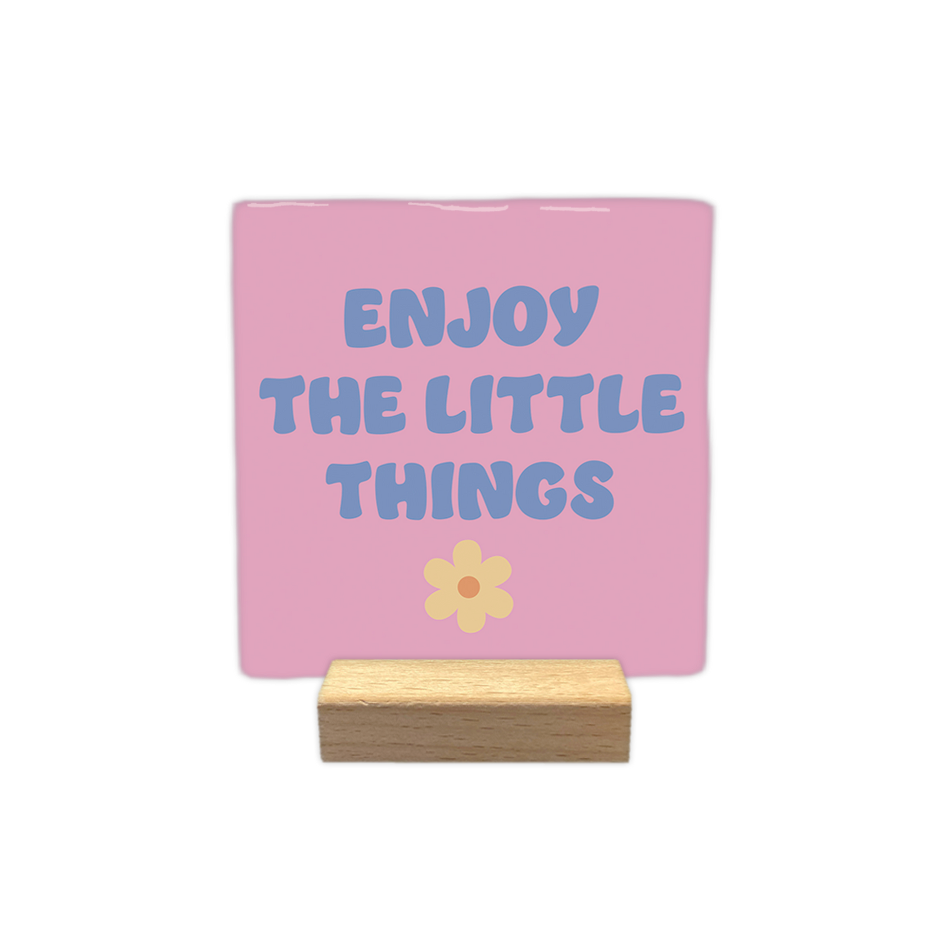 Enjoy the little things - Decoratie tegel - Formaat: - Op deze decoratietegel staat in het midden de Engelse tekst "ENJOY THE LITTLE THINGS". De tegel zelf is roze en vierkant, en staat in een houten standaard. De tekst is verdeeld over drie regels en geschreven in een speels, blauwachtig paars lettertype in hoofdletters, met daaronder een kleine, lichtgele bloem met een oranje rondje. - Snelwenskaart