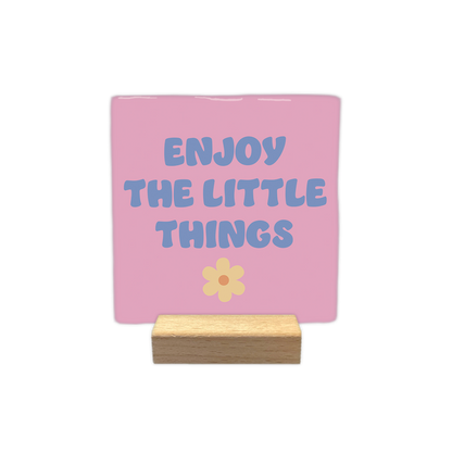 Enjoy the little things - Decoratie tegel - Formaat: - Op deze decoratietegel staat in het midden de Engelse tekst "ENJOY THE LITTLE THINGS". De tegel zelf is roze en vierkant, en staat in een houten standaard. De tekst is verdeeld over drie regels en geschreven in een speels, blauwachtig paars lettertype in hoofdletters, met daaronder een kleine, lichtgele bloem met een oranje rondje. - Snelwenskaart