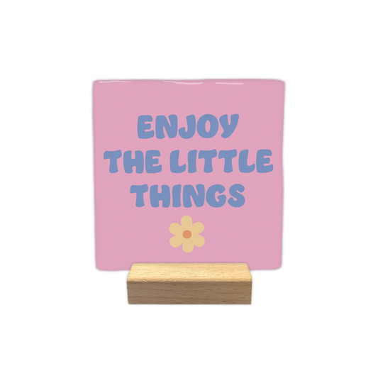 Enjoy the little things - Decoratie tegel - Formaat: - Op deze decoratietegel staat in het midden de Engelse tekst "ENJOY THE LITTLE THINGS". De tegel zelf is roze en vierkant, en staat in een houten standaard. De tekst is verdeeld over drie regels en geschreven in een speels, blauwachtig paars lettertype in hoofdletters, met daaronder een kleine, lichtgele bloem met een oranje rondje. - Snelwenskaart