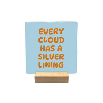 Every cloud has a silver lining - Decoratie tegel - Formaat: - Op deze decoratietegel staat de tekst "EVERY CLOUD HAS A SILVER LINING" in hoofdletters gedrukt. De tekst is uitgevoerd in een oranje kleur en is gecentreerd op een lichtblauwe, vierkante achtergrond. Het lettertype heeft een speelse, afgeronde stijl. De tegel heeft een gladde, glanzende afwerking. - Snelwenskaart