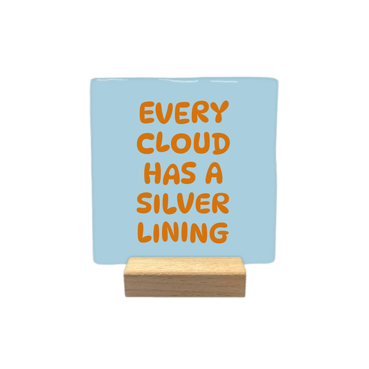 Every cloud has a silver lining - Decoratie tegel - Formaat: - Op deze decoratietegel staat de tekst "EVERY CLOUD HAS A SILVER LINING" in hoofdletters gedrukt. De tekst is uitgevoerd in een oranje kleur en is gecentreerd op een lichtblauwe, vierkante achtergrond. Het lettertype heeft een speelse, afgeronde stijl. De tegel heeft een gladde, glanzende afwerking. - Snelwenskaart
