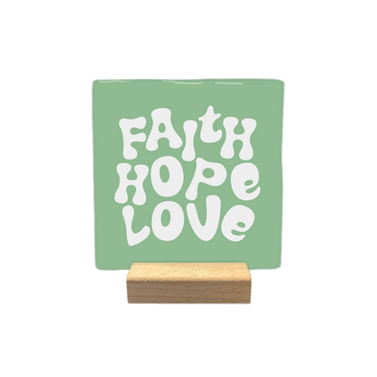 Faith hope love - Decoratie tegel - Formaat: - Op deze decoratietegel staat de tekst "FAITH HOPE LOVE" in witte letters met een speels, golvend lettertype. De tekst is gecentreerd op een groene, vierkante achtergrond. - Snelwenskaart