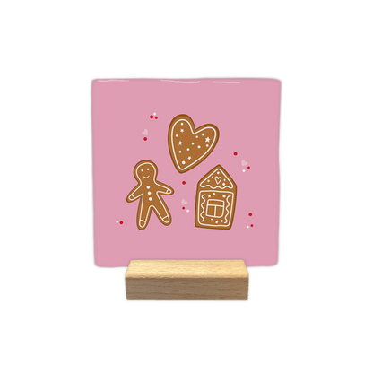 Gingerbread - Decoratie tegel - Op deze decoratietegel staat een illustratie van drie peperkoekfiguren tegen een lichtroze achtergrond: een hart, een peperkoekmannetje en een peperkoekhuisje, alle voorzien van witte decoratieve details. Rondom de figuren zijn kleine rode stippen en roze hartjes afgebeeld. - Snelwenskaart