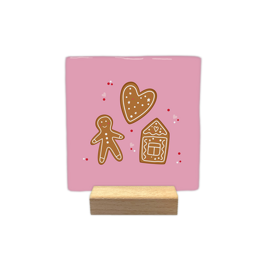 Gingerbread - Decoratie tegel - Op deze decoratietegel staat een illustratie van drie peperkoekfiguren tegen een lichtroze achtergrond: een hart, een peperkoekmannetje en een peperkoekhuisje, alle voorzien van witte decoratieve details. Rondom de figuren zijn kleine rode stippen en roze hartjes afgebeeld. - Snelwenskaart
