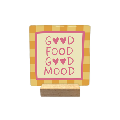 Good food good mood - Decoratie tegel - Formaat: - Op deze decoratietegel staat de tekst "GOOD FOOD GOOD MOOD" in roze letters, waarbij de letter 'O' in de woorden 'GOOD' is vervangen door roze hartjes. De tekst is geplaatst op een crèmekleurig middenveld dat wordt omlijst door een roze rand. De achtergrond van de tegel heeft een oranje-geel ruitpatroon. - Snelwenskaart
