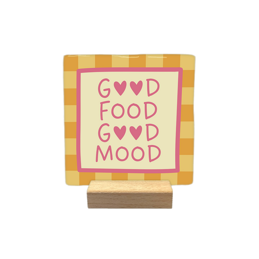 Good food good mood - Decoratie tegel - Formaat: - Op deze decoratietegel staat de tekst "GOOD FOOD GOOD MOOD" in roze letters, waarbij de letter 'O' in de woorden 'GOOD' is vervangen door roze hartjes. De tekst is geplaatst op een crèmekleurig middenveld dat wordt omlijst door een roze rand. De achtergrond van de tegel heeft een oranje-geel ruitpatroon. - Snelwenskaart