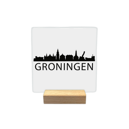 Groningen skyline - Decoratie tegel - Formaat: - Op deze decoratie tegel staat een zwarte skyline van Groningen afgebeeld tegen een witte achtergrond. Het silhouet toont diverse herkenbare gebouwen uit de stad, waaronder de Martinitoren. Onder de skyline staat in zwarte letters de plaatsnaam Groningen. Het gehele ontwerp is uitgevoerd in een minimalistische zwart-witstijl. - Snelwenskaart