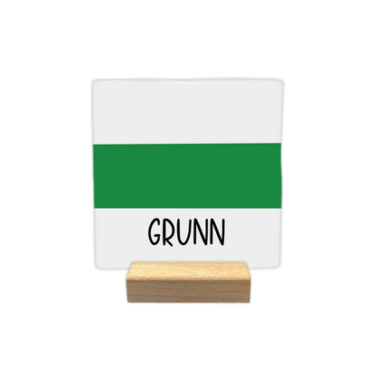 Grunn - Decoratie tegel - Formaat: - Op deze decoratie tegel staat de tekst "GRUNN" in zwarte letters aan de onderzijde. De achtergrond bestaat uit een horizontale groene baan in het midden, omringd door witte vlakken boven en onder. - Snelwenskaart
