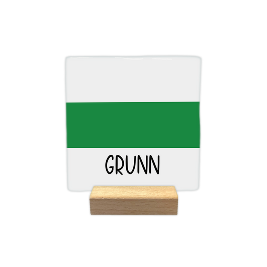 Grunn - Decoratie tegel - Formaat: - Op deze decoratie tegel staat de tekst "GRUNN" in zwarte letters aan de onderzijde. De achtergrond bestaat uit een horizontale groene baan in het midden, omringd door witte vlakken boven en onder. - Snelwenskaart