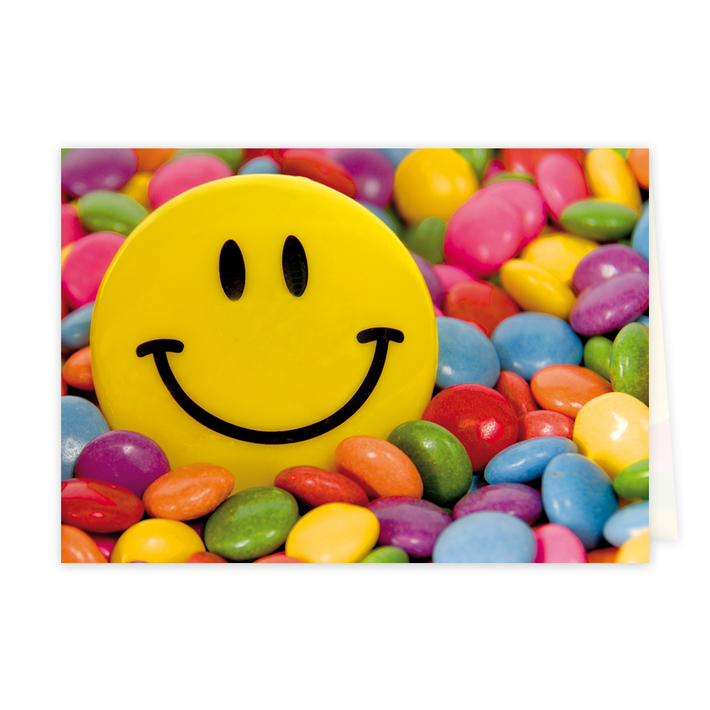 Op deze wenskaart staat een vrolijke afbeelding van een grote gele smiley die omringd wordt door een overvloed aan kleurrijke suikerbolletjes, vergelijkbaar met Smarties of M&M's. De suikerbolletjes hebben diverse felle kleuren zoals roze, blauw, groen, oranje, paars en geel, en vullen de gehele achtergrond en voorgrond rondom de smiley. De smiley zelf heeft het klassieke ontwerp met twee zwarte ovalen als ogen en een brede zwarte glimlach.