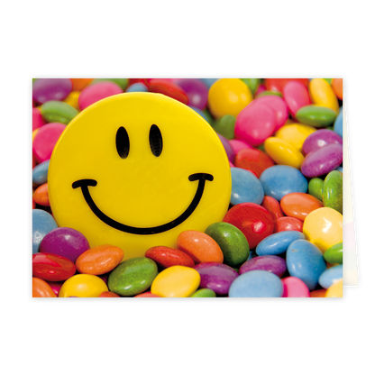 Op deze wenskaart staat een vrolijke afbeelding van een grote gele smiley die omringd wordt door een overvloed aan kleurrijke suikerbolletjes, vergelijkbaar met Smarties of M&M's. De suikerbolletjes hebben diverse felle kleuren zoals roze, blauw, groen, oranje, paars en geel, en vullen de gehele achtergrond en voorgrond rondom de smiley. De smiley zelf heeft het klassieke ontwerp met twee zwarte ovalen als ogen en een brede zwarte glimlach.