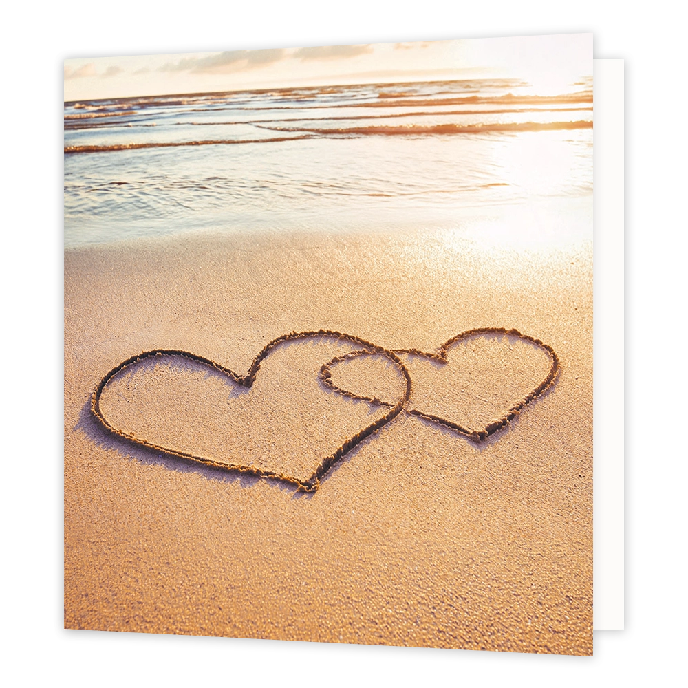 Hartjes op het strand - Formaat: Standaard - Vorm: Vierkant - Blanco kaart - Op deze wenskaart staat een illustratie van twee in elkaar geschoven harten die in het natte zand op een strand zijn getekend. De achtergrond toont de zee en het strand tijdens een warme zonsondergang of zonsopgang. Er is geen tekst op de illustratie zichtbaar. - Snelwenskaart