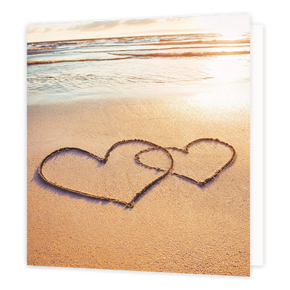 Hartjes op het strand - Formaat: Standaard - Vorm: Vierkant - Blanco kaart - Op deze wenskaart staat een illustratie van twee in elkaar geschoven harten die in het natte zand op een strand zijn getekend. De achtergrond toont de zee en het strand tijdens een warme zonsondergang of zonsopgang. Er is geen tekst op de illustratie zichtbaar. - Snelwenskaart