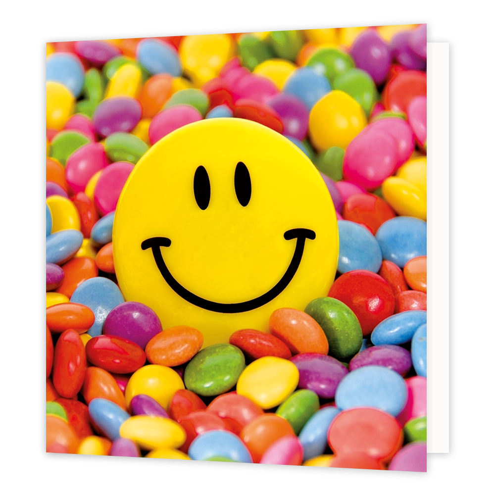 Smarties - Formaat: Standaard - Vorm: Vierkant - Blanco kaart - Op deze wenskaart staat een illustratie van een grote, gele smileyknop centraal afgebeeld, omringd door een kleurrijke zee van kleine, gekleurde snoepjes in diverse tinten zoals rood, blauw, groen en paars. De smiley heeft twee zwarte ovale ogen en een brede, zwarte glimlach. Er is geen tekst op de illustratie zichtbaar. - Snelwenskaart